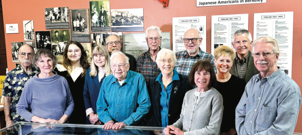 A group of thirteen individuals posing together in front of a display about Japanese Americans in Berkeley, with historical photographs and informational panels in the background.