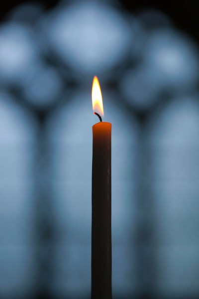A lit candle with a flickering flame in front of a blurred background resembling stained glass.